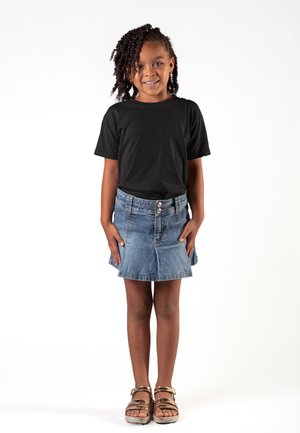 Ragazza giovane con capelli ricci che indossa una t-shirt nera, gonna di jeans e sandali, in piedi e sorridente contro uno sfondo bianco.