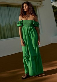 Robe maxi verte sans épaule avec des manches volantes, buste smocké, jupe fluide et ourlet à volants ; tissu en coton léger.