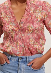 Floral button-up blouse in pink with ruffles and pleats, featuring a mix of yellow and green floral patterns. Paired with blue denim jeans.
