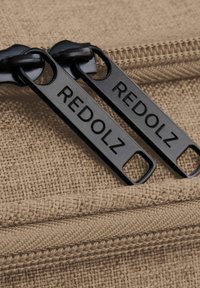 Tiranti della cerniera neri con la scritta "REDOLZ" incisa, attaccati a un tessuto beige testurizzato, che mostra un aspetto intrecciato e una traccia della cerniera visibile.