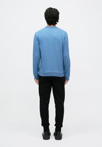 Μπλε φούτερ μακρυμάνικο με ριμπ μανσέτες και τρέσα, φορεμένο με μαύρο αθλητικό παντελόνι και μαύρα παπούτσια. Ομαλή υφή, μινιμαλιστικός σχεδιασμός.