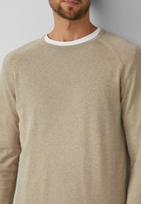 Hellbeiger Pullover aus weichem Stoff, mit einem Rundhalsausschnitt, der mit einem weißen Innenfutter versehen ist, und langen Raglanärmeln.