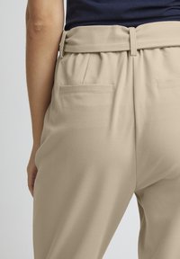 Beige Hosen mit glatter Textur, hoher Taille und einem Gürtel-Design sowie zwei Gesäßtaschen. Der Stoff wirkt leicht und fließend.