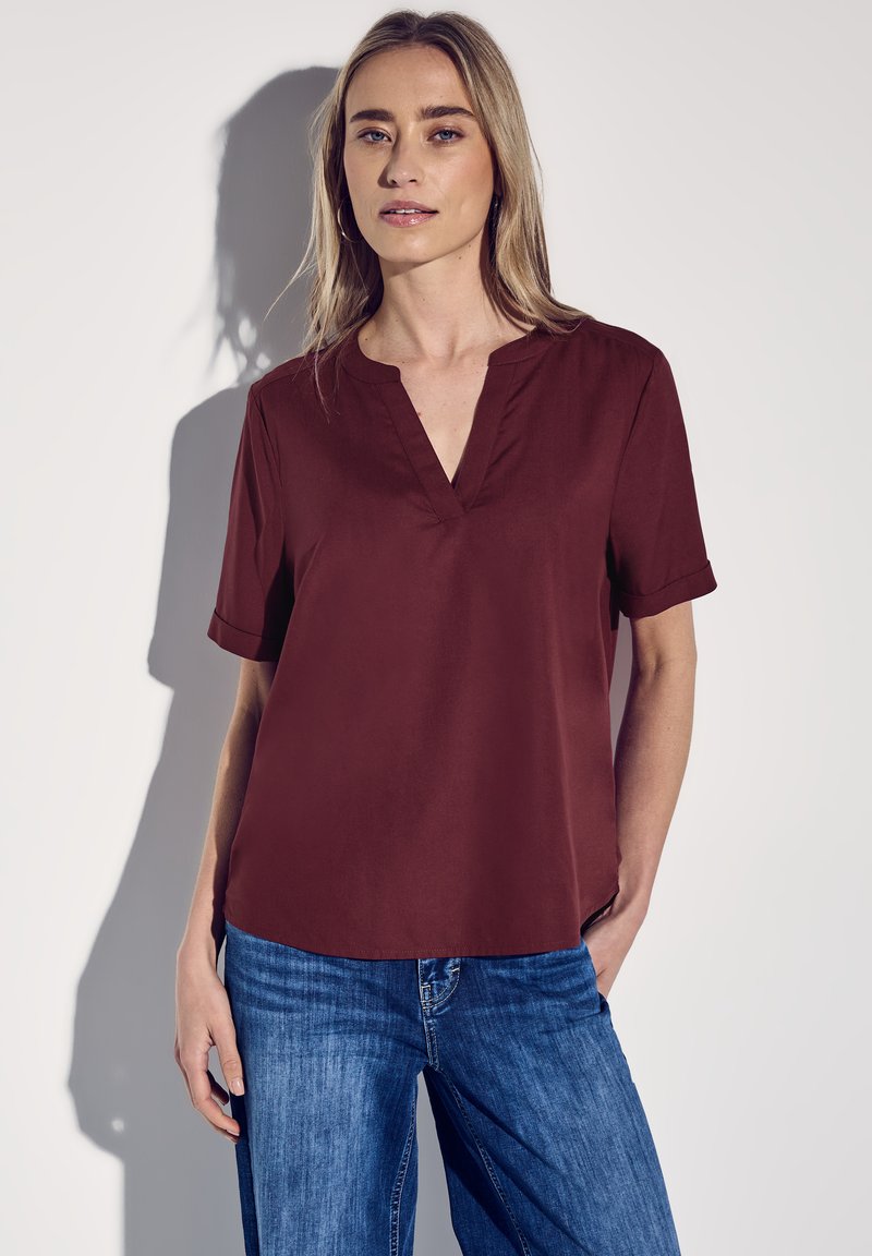 Bordeaux kortärmad blus med V-ringning och uppvikta ärmar, som är ihopparad med blå denimjeans. Slät textur och avslappnad passform.