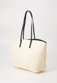 Sac fourre-tout en toile crème avec de longues anses en cuir noir et une base rectangulaire, présenté sur un fond blanc.