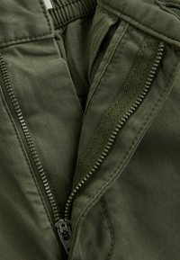 Pantalon vert olive avec une fermeture éclair et un tissu texturé. Matière lisse et durable avec une ceinture élastique et des poches latérales.