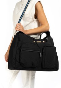 Black nylon tote bag with two front pockets, padded texture, silver hardware, and a multicolored braided shoulder strap.