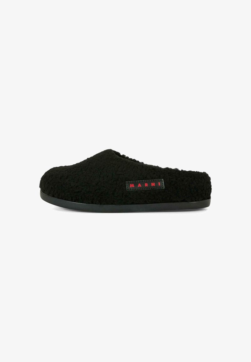 Chausson noir avec un extérieur texturé et duveteux, et une semelle plate noire. Présente une petite étiquette logo rouge sur le côté.