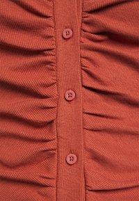 Rostfärgad topp med ribbad textur, med samlad tyg och fyra knappstängning. Knapparna är rosa med en tvåhålsdesign.