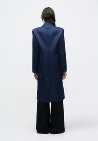 Tmavomodrý dlhý kabát s konštrukčne tvarovanými ramenami, výrezovým golierom a zapínaním na jeden gombík. Má dlhé rukávy a priliehavý strih na zadnej strane.
