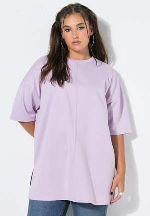 Ung kvinde med langt mørkt hår og store hoop-øreringe iført en løs lys lilla T-shirt og blå jeans mod en hvid baggrund.