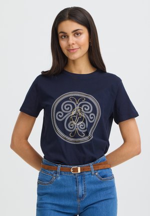 Femme aux longs cheveux foncés portant un T-shirt bleu marine avec un motif géométrique circulaire, un jean bleu et une ceinture marron, debout devant un fond uni.