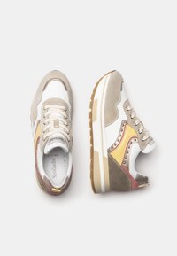 Zapatillas beige y blancas con texturas de ante y malla, acentuadas por detalles en amarillo y rojo, con tachuelas de metal y una suela de goma gruesa.