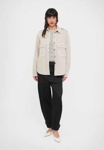 Μπεζ πουκάμισο-σακάκι με κουμπιά και δύο τσέπες μπροστά, φορεμένο πάνω από ριγέ τοπ, συνδυασμένο με μαύρο παντελόνι με φαρδιά πόδια και λευκές μπαλαρίνες.