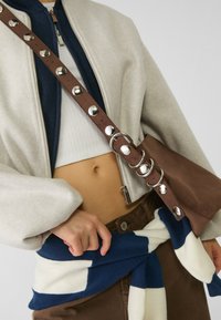 Brun lædertaske med sølv detaljer, båret crossbody over en creme bomberjakke og en kort hvid top, mod et blå- og hvidstribet tørklæde.