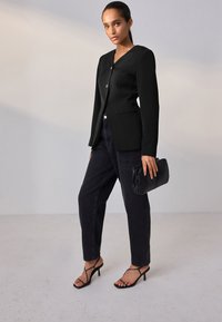 Blazer nero con scollo a V, chiusura con un solo bottone e maniche lunghe; abbinato a jeans scuri e décolleté nere con cinturini, con una pochette in mano.
