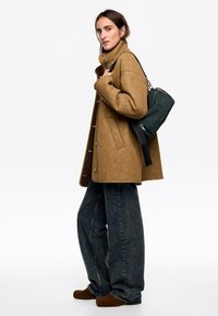 Abrigo de cuero sintético marrón con cuello alto y botones, combinado con jeans de mezclilla oscuros de pierna ancha y botas de ante marrones; llevando un bolso verde.