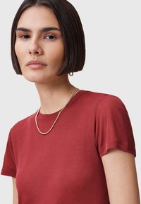 Czerwony t-shirt z krótkim rękawem, wykonany z gładkiego, lekkiego materiału. Posiada okrągły dekolt. Modelka nosi cienki złoty naszyjnik jako akcent.