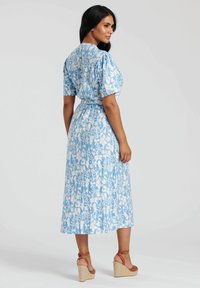 Niebieska sukienka w kwiatowy wzór z krótkimi bufkowymi rękawami, wcięciem w talii i długości midi, zestawiona z brązowymi sandałami na koturnie.
