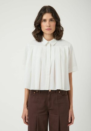 Kvinne med skulderlangt brunt hår iført hvit kortermet plissert bluse og høytlivs mørkebrune bukser, står mot hvit bakgrunn.