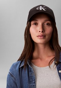 Schwarze Baseballmütze mit weißem "CK"-Logo vorne, getragen über braunem Haar. Blaue Jeansjacke mit sichtbar darunterliegendem hellgrauen Hemd.