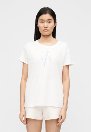 Witte katoenen T-shirt met korte mouwen, met een lichtblauw grafisch "A/X" ontwerp centraal op de borst. Gecombineerd met lichte shorts.