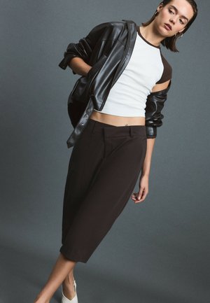Femme portant une veste en cuir noire tombant des épaules, un haut sans manches blanc et noir, un pantalon large noir raccourci et des chaussures blanches sur un fond uni.