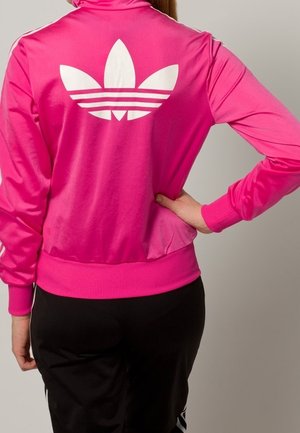 Vaaleanpunainen urheilutakki, jossa on korkea kaulus ja suuri valkoinen kolmen lehden logo selässä sekä resorit hihansuissa ja helmassa. Valmistettu kiiltävästä materiaalista.