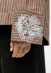 Brown striped shirt with white floral embroidery on the cuff. Cotton material; fitted style; smooth texture. Cuff detail prominent.