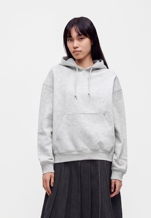 Femme aux longs cheveux foncés portant un sweat à capuche gris clair et une jupe plissée foncée, debout devant un fond blanc uni.