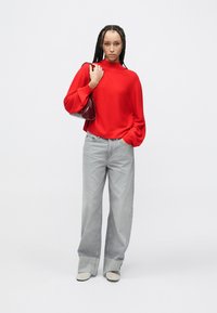 Czerwony sweter z długimi, bufiastymi rękawami, zestawiony z jasnoszarymi spodniami o szerokich nogawkach. Modelka trzyma małą bordową torebkę.