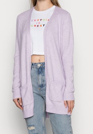 Cardigan en tricot lavande avec un devant ouvert, des poignets côtelés et deux poches avant, porté sur un t-shirt blanc court et un jean bleu déchiré.