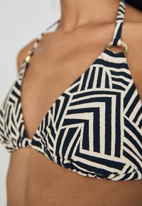 Bikini topp med trekantet design, svart og hvit geometrisk mønster, gullfarge detaljer, og justerbare stropper.