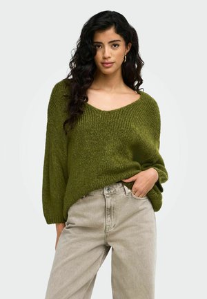 Femme aux longs cheveux bouclés porte un pull ample vert olive et un jean beige clair taille haute, posant avec une main dans la poche.
