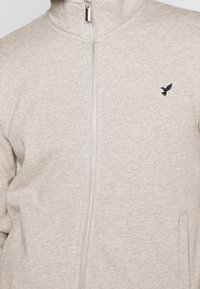 Chaqueta beige con cremallera y cuello alto, con un logo de pájaro bordado en negro en el lado izquierdo del pecho.
