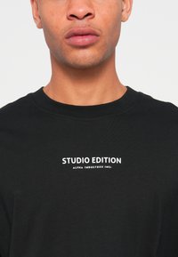 Juoda medvilninė marškinėlių su apvalia apykakle, trumpomis rankovėmis, priekyje su baltais užrašais "STUDIO EDITION ALPHA INDUSTRIES INC."