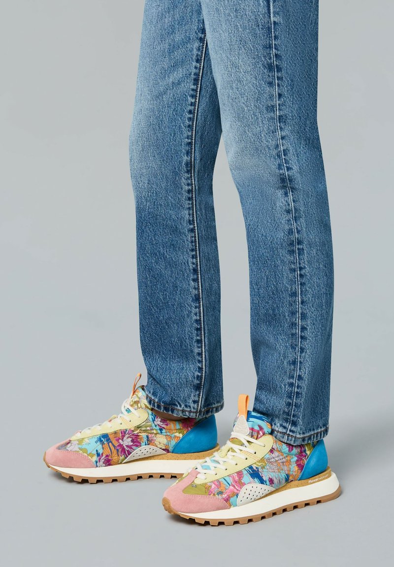 Baskets multicolores avec des motifs floraux, accents en daim rose et bleu, lacets jaunes et une semelle texturée, associées à un jean en denim bleu.