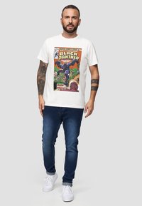 Vit bomullströja med en färgglad grafisk tryck av Black Panther-serietidningens omslag. Kombinerad med mörkblå jeans och vita sneakers.