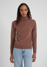 Brauner Pullover mit Mock-Neck aus weichem Stoff, mit langen Ärmeln und einem enganliegenden Taillenbund. Kombiniert mit hellblauen Jeans.