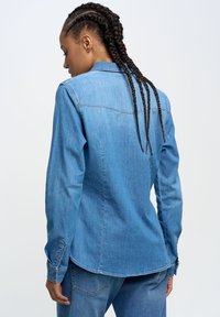 Denimhemd in Hellblau, mit einem Button-Down-Kragen, langen Ärmeln und einem abgerundeten Saum. Die Rückansicht zeigt Nahtdetails und einen strukturierten Stoff.