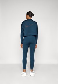 Tamno plavi skraćeni dukserica s crnim Nike logotipom na leđima, u kombinaciji s odgovarajucim tamnoplave tajicama koje imaju mali plavi Nike logotip na gležnju.