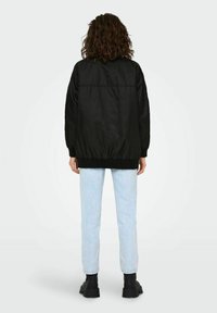 Veste bomber légère noire avec un ourlet côtelé, présentant une texture lisse et une coupe décontractée, associée à un jean droit bleu clair et des bottes noires.