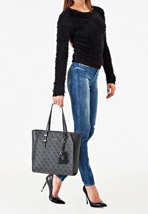Femme portant un pull noir en maille pelucheuse, un jean skinny bleu, des talons hauts noirs, tenant un grand sac fourre-tout noir à motifs par les anses.