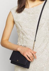 Svart texturerad läder crossover-väska med ett guldkedjeslang och en subtil logotyp. Har en klaffstängning och rektangulär form.