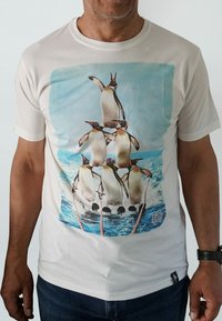 Crème-kleurig katoen t-shirt met een afbeelding van vijf pinguïns die op elkaar balanceren tegen een blauwe oceaan achtergrond.
