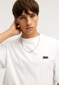 Hvid bomulds t-shirt med et sort broderet logo på venstre bryst, parret med et sølvmetal kæde halskæde, glat tekstur.