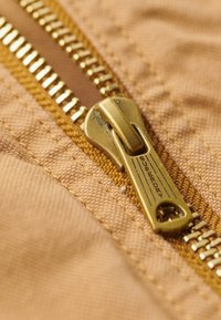 Fermeture éclair en métal doré sur un tissu beige avec une finition texturée et une couture contrastante, dotée d'un langot de marque et de dents visibles.