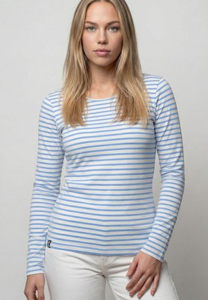 Junge Frau mit langen blonden Haaren, die ein blau-weiß gestreiftes Langarmshirt und weiße Hose trägt und vor einem schlichten grauen Hintergrund steht.