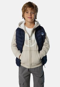 Chaleco acolchado navy sobre una sudadera beige clara con logo. Pantalones cargo gris claro. Textura suave, frente con cremallera, look casual en capas.