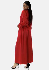 Robe maxi rouge avec manches longues bouffantes, taille cintrée et jupe fluide. Portée avec des sandales à talons hauts noires, tissu lisse et détails minimaux.